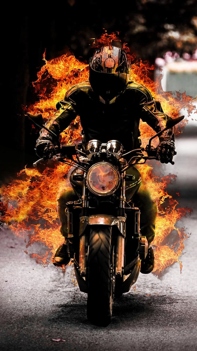 Motorcyclist in a leather jacket riding a classic bike engulfed in bright orange flames at a low angle.