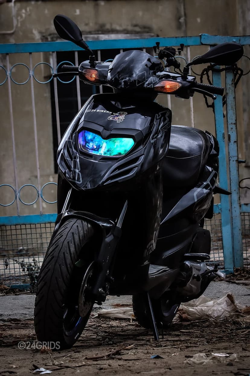 Black motor scooter with glowing blue and green LED headlights parked near a blue metal fence on gritty ground.