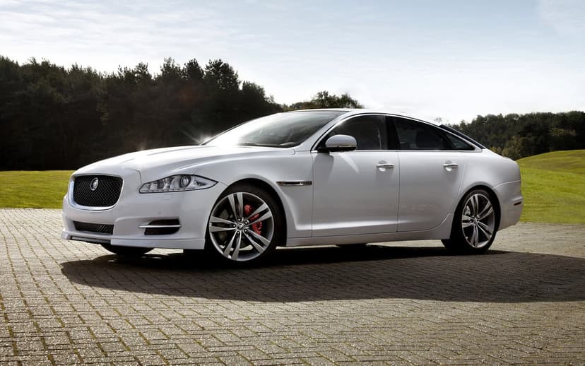 White Jaguar XJ sedan on a stone-paved driveway with green hills and a forest under a bright sky.