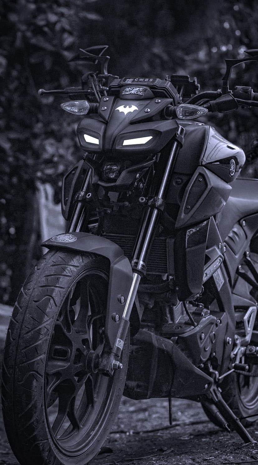 Matte black motorcycle front with a bat logo and glowing white LED headlights against a dark blurred background.