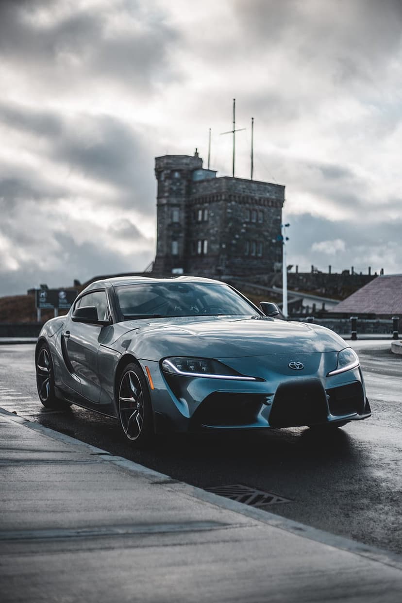 Grey Toyota Supra on rain-slicked asphalt before a historic stone tower under a cloudy, overcast, moody sky.