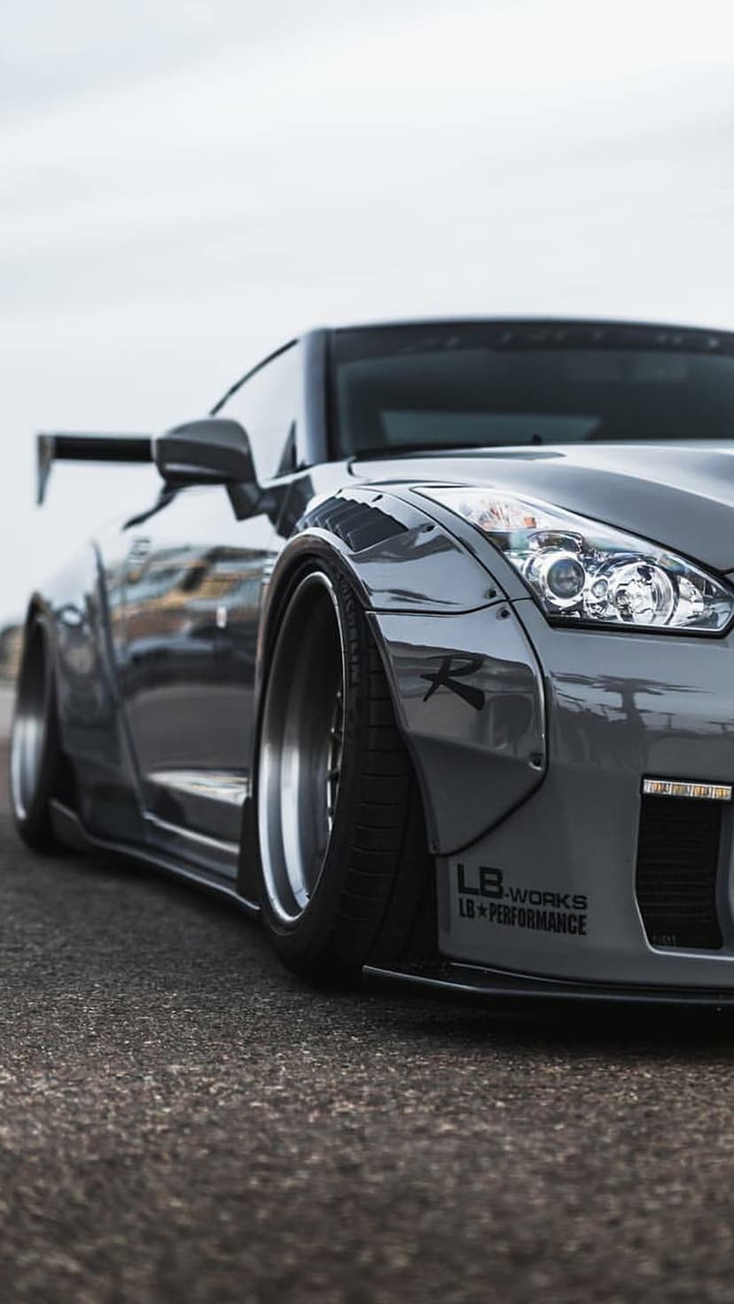 Nissan GT-R featuring bolted widebody fenders and LB-Works decals sits low on dark asphalt against a grey sky.
