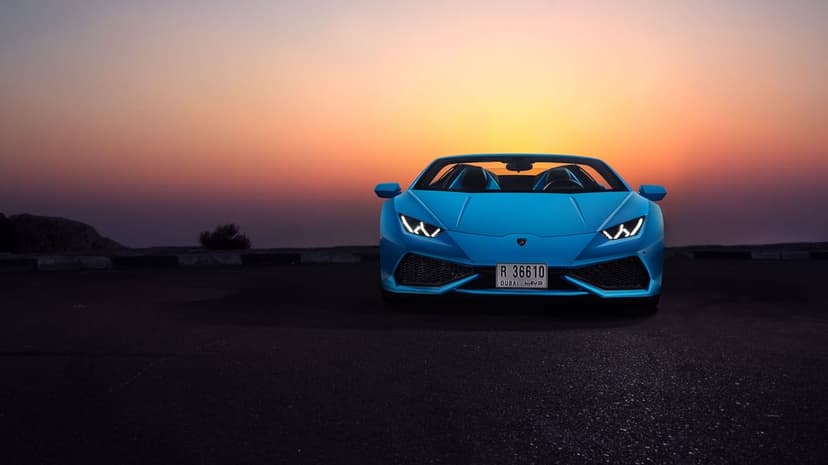 Blue Lamborghini Huracán Spyder convertible at sunset with glowing headlights on a dark asphalt road.