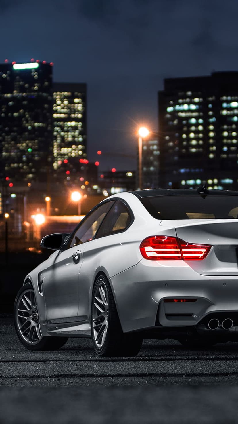 White BMW M4 coupe parked on asphalt at night with glowing taillights and a bokeh city skyline background.