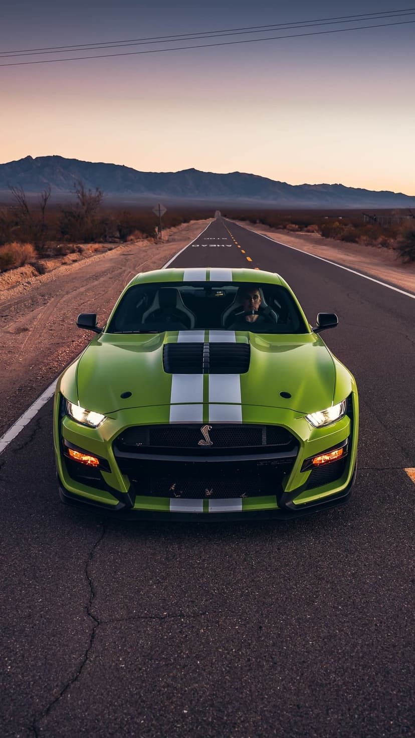 Front view of a lime green Shelby GT500 with white stripes on a desert road under a sunset sky.