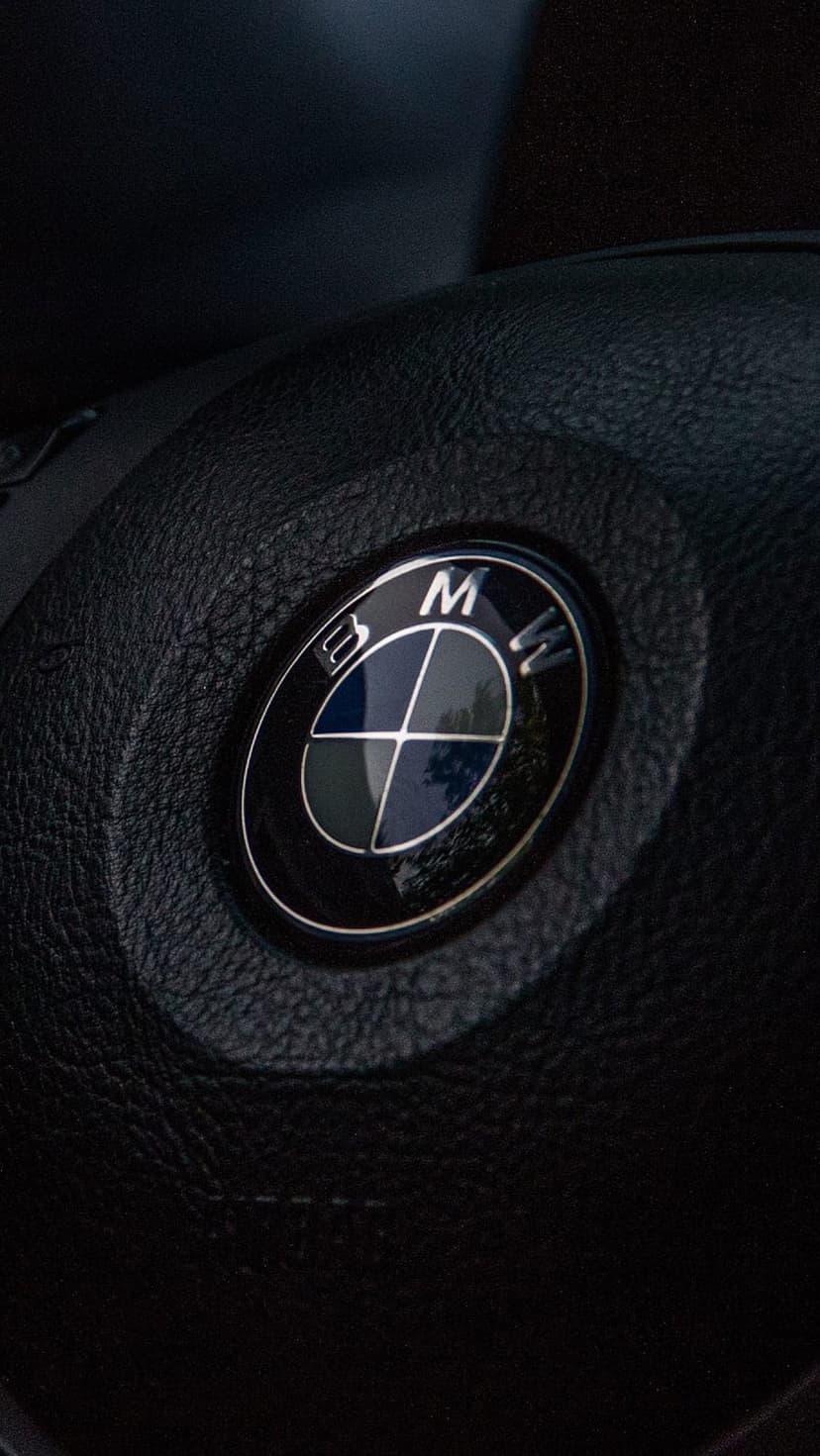 BMW emblem in blue and white centered on a black leather steering wheel with polished metallic reflections.