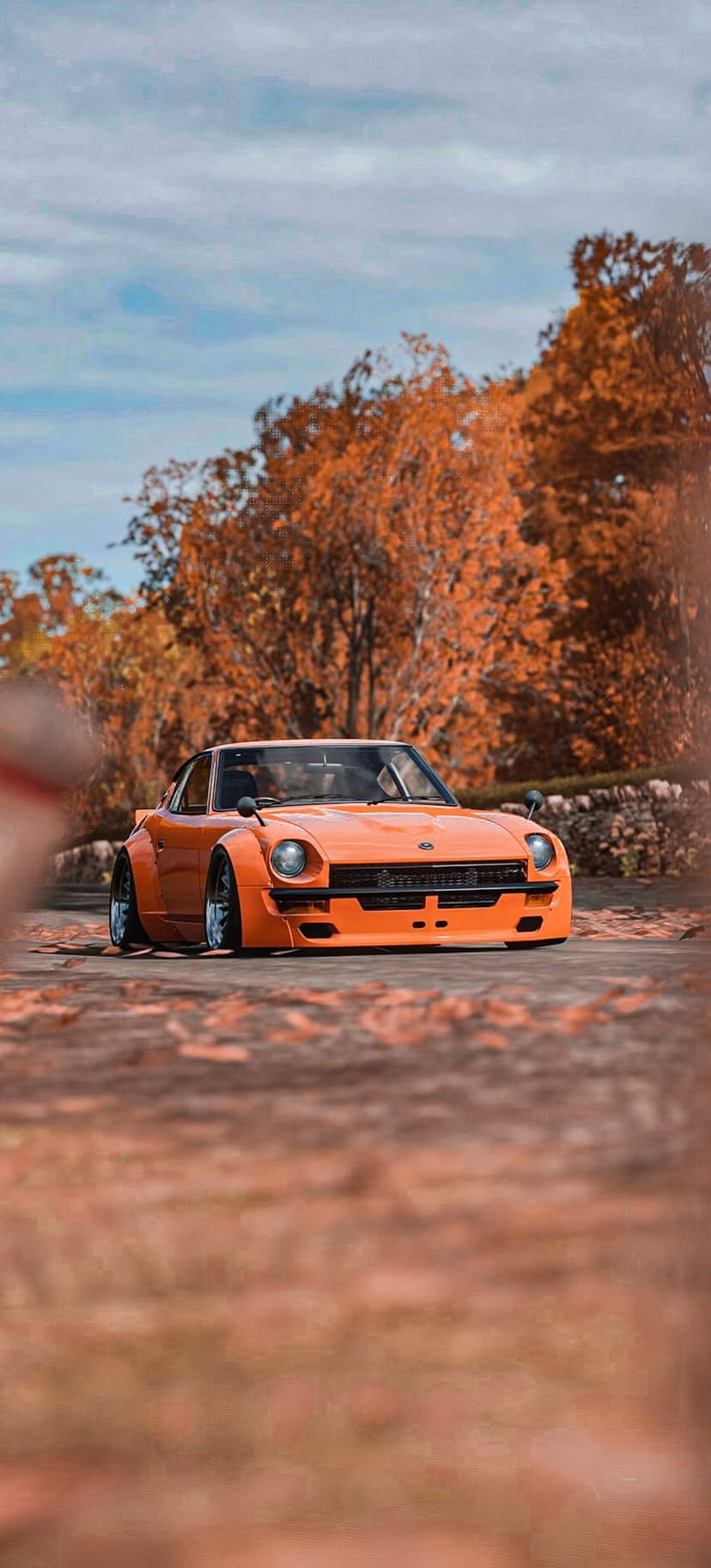 Orange Nissan Fairlady Z S30 with wide fenders parked on a road covered in red autumn leaves under a cloudy sky.