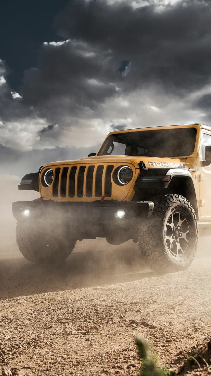 Yellow Jeep Rubicon off-roading through a cloud of dust under dark dramatic clouds on a sandy desert road.