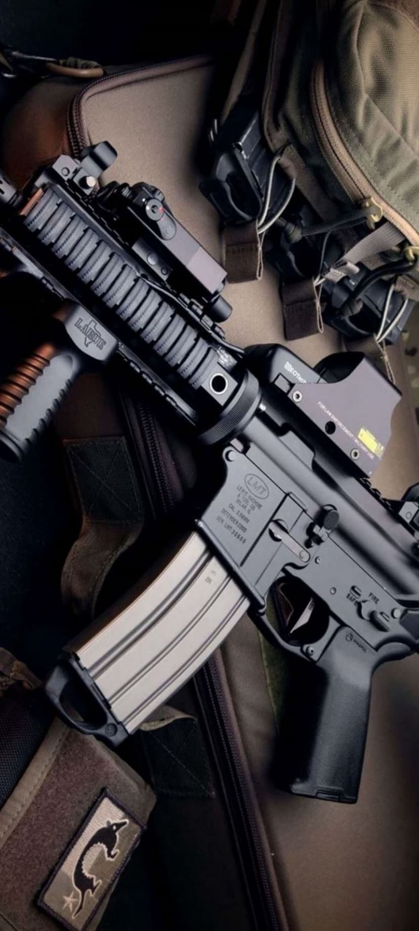Tactical rifle with a red dot sight and vertical foregrip rests on a tan backpack and equipment pouches.