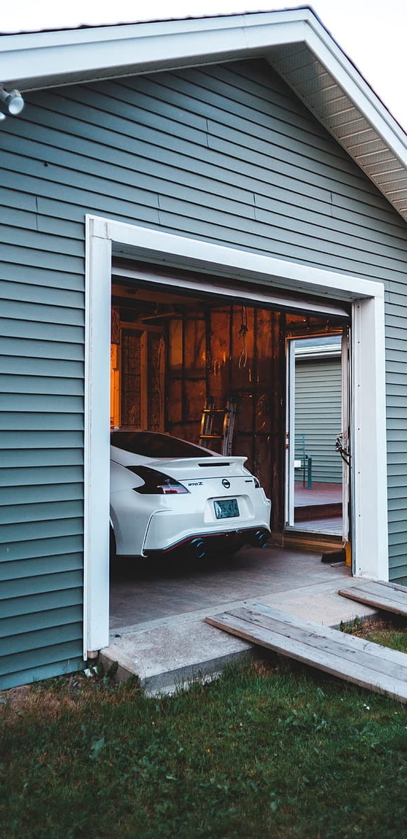 White Nissan 370Z sports car rear with quad exhausts parked inside a rustic blue wooden garage during dusk.