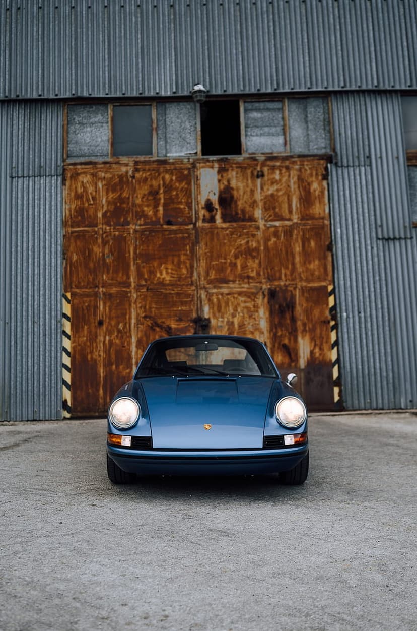 Blue Porsche 911 head-on in front of rusty corrugated metal doors on a weathered concrete industrial floor.