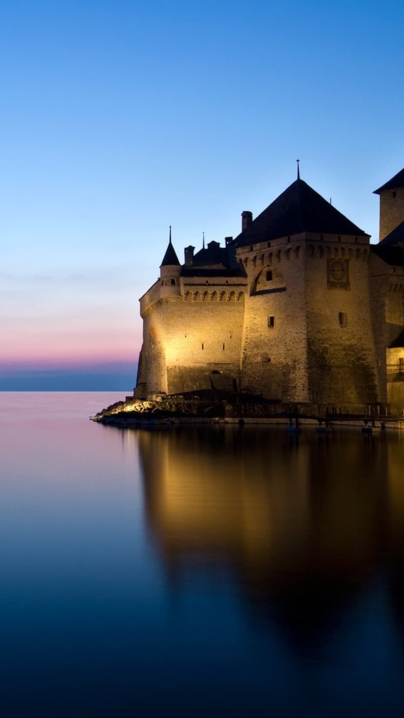 Château de Chillon fortress illuminates the shore of Lake Geneva under a purple and blue twilight sky.