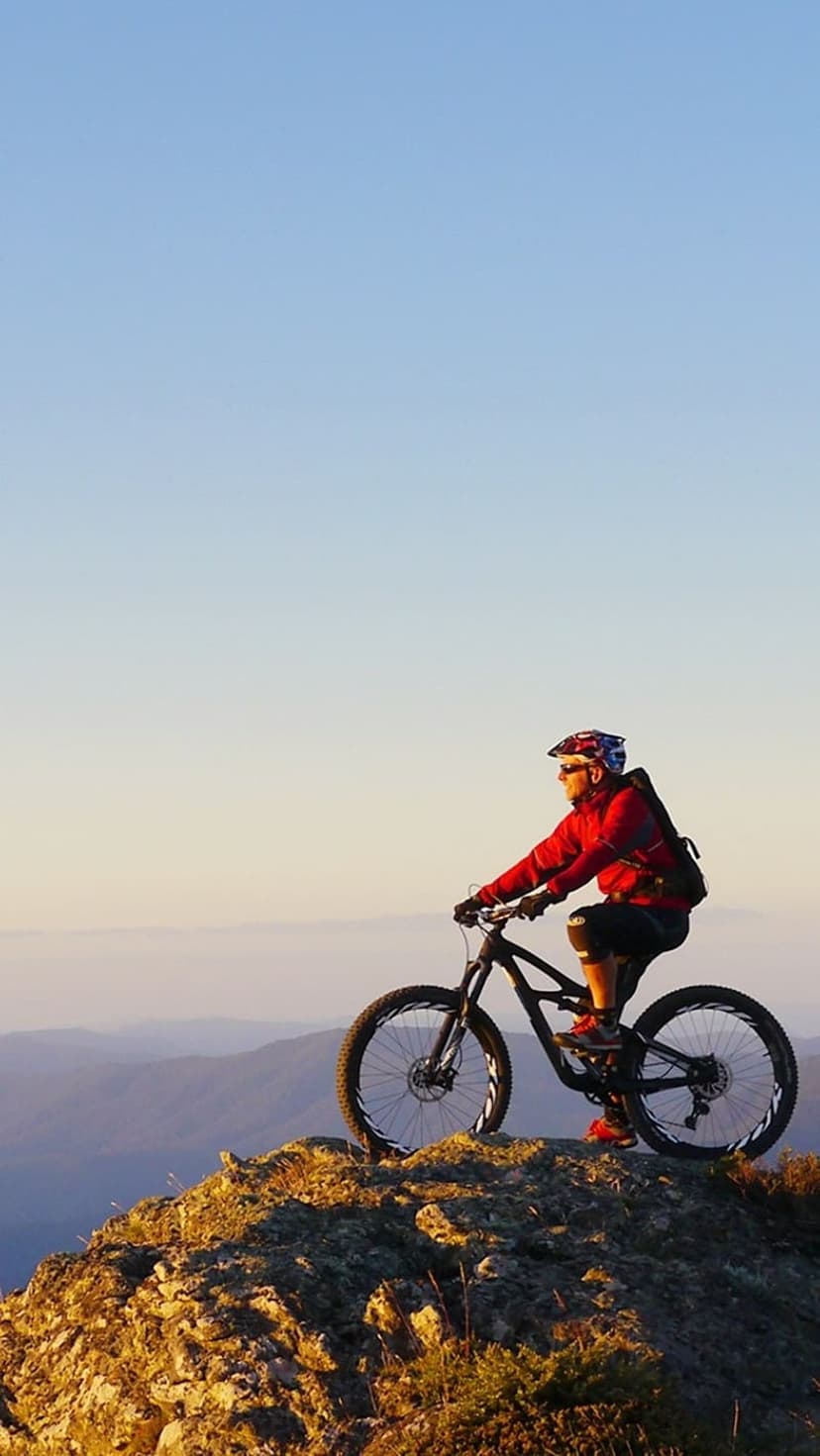 Mountain biker in a red jacket rests on a rocky peak overlooking distant blue mountains during a sunset silhouette.