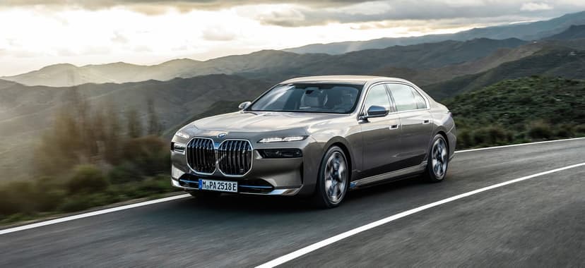 BMW i7 luxury sedan driving on a mountain road with motion-blurred wheels under a hazy sunset sky.
