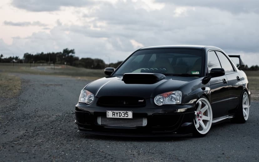 Black Subaru WRX STi blobeye with white wheels and front intercooler parked on a road under a cloudy sky.