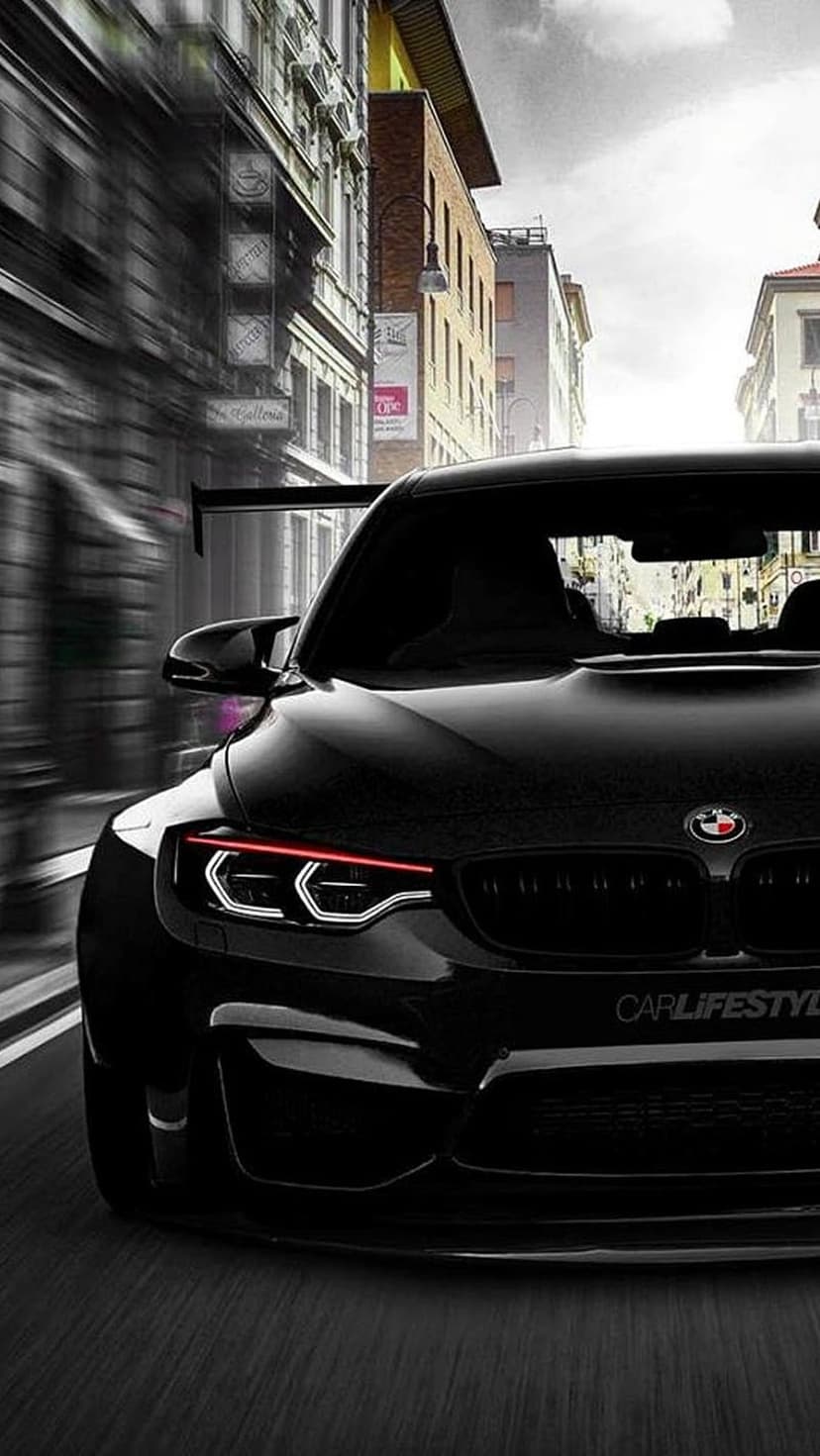 Black BMW M4 sports car with glowing headlights driving through a blurred European city street at night.