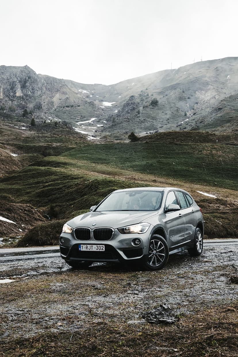 Silver BMW X1 SUV parked on a wet gravel mountain road with snow-covered peaks under a misty, overcast alpine sky.