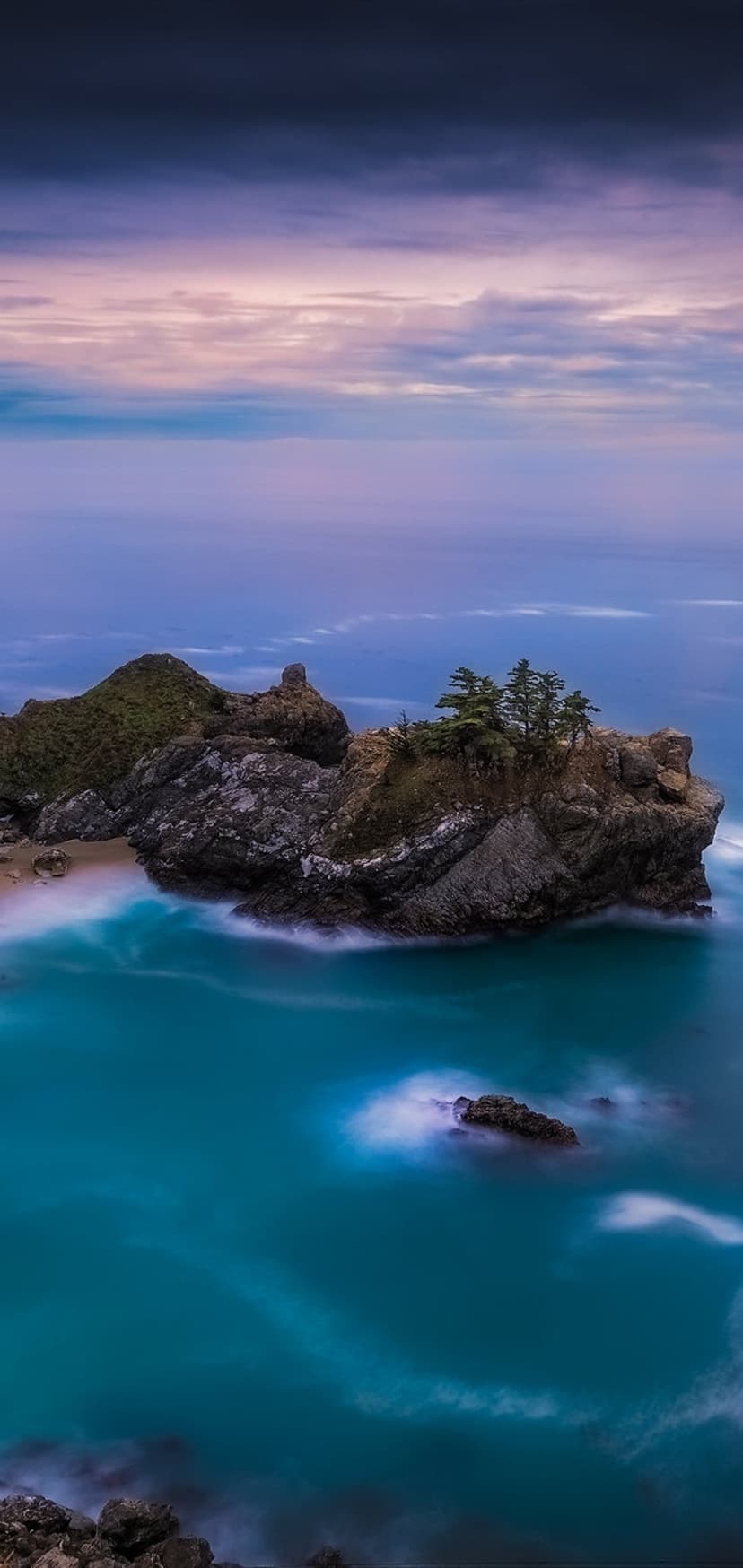 Emerald waters lap against rocky cliffs and a secluded beach under a purple and pink dawn sky in a vertical view.