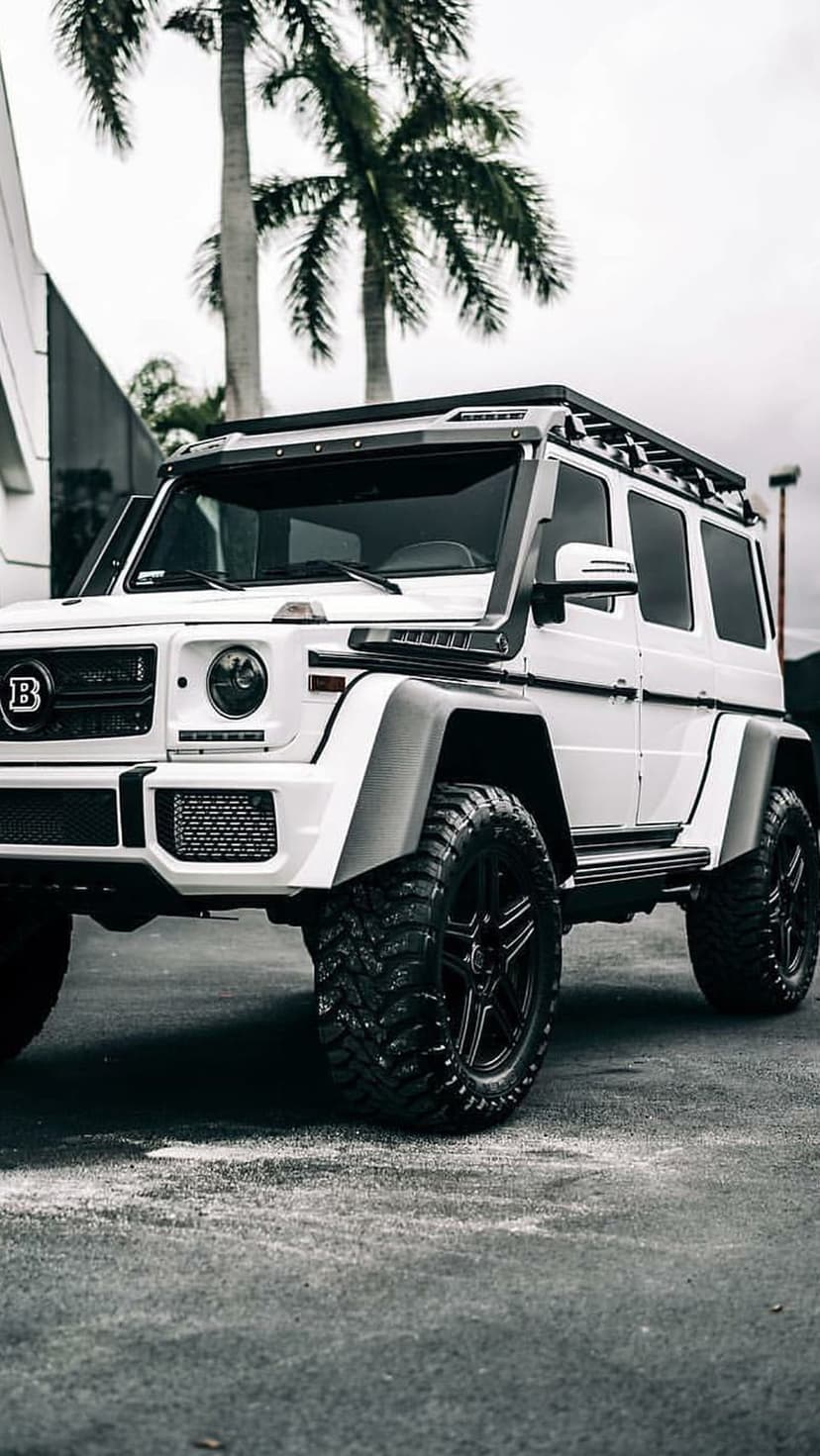 White Mercedes G-Wagon with oversized black tires, roof lights, and a Brabus logo parked near palm trees.