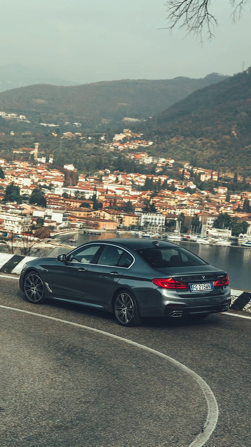 BMW sedan drives on a winding hillside road overlooking a Mediterranean village with boats and terracotta roofs.