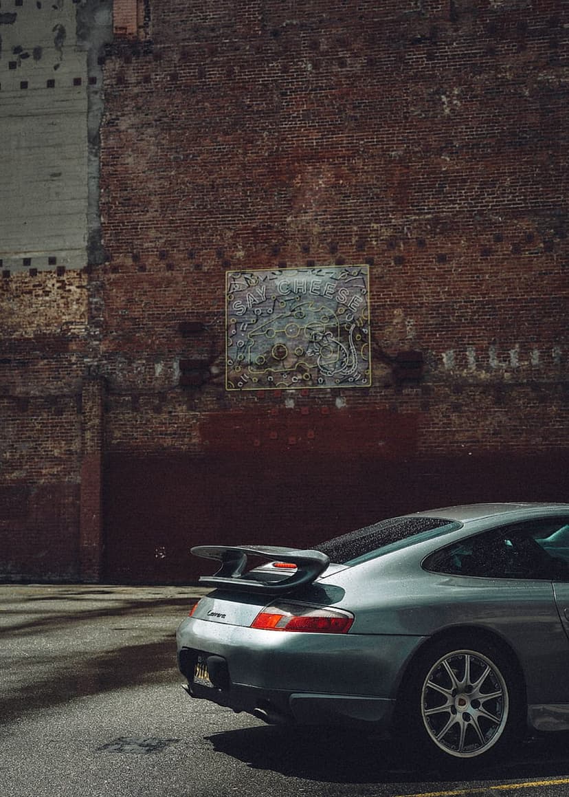 Silver Porsche 911 GT3 with a large rear wing parked beside a brick wall featuring framed abstract street art.