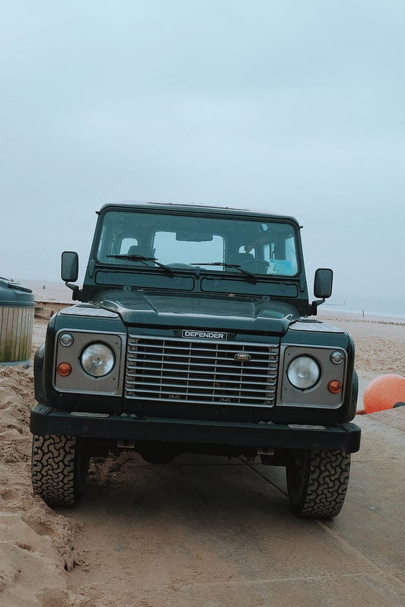 Dark green Land Rover Defender parked on a sandy beach under a moody overcast sky with rugged off-road tires.