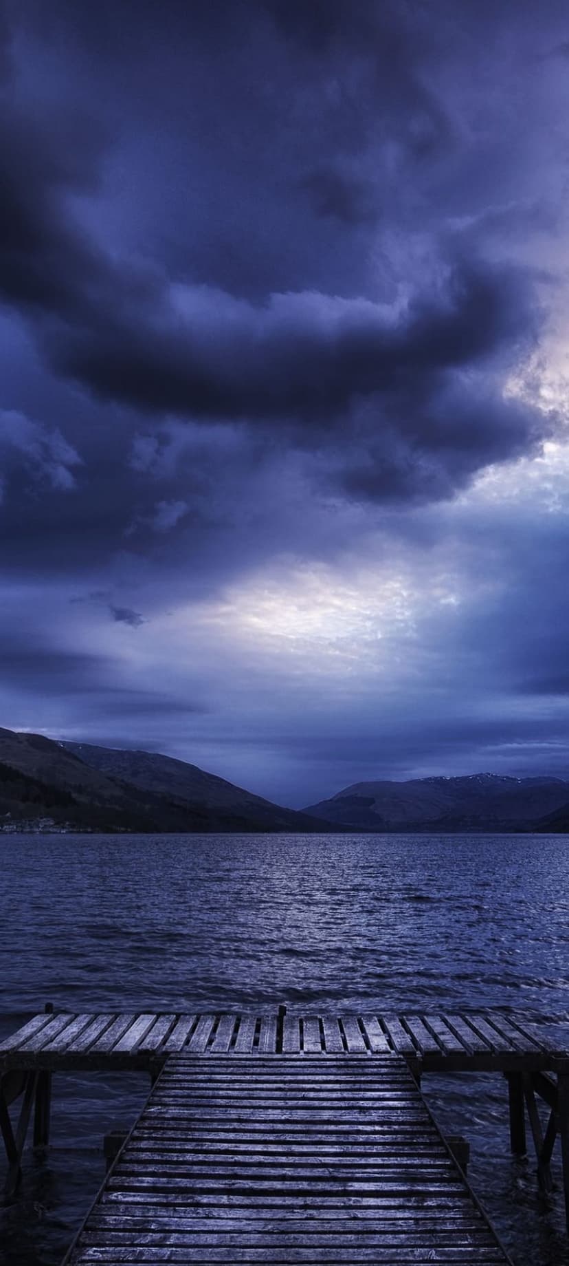 Wooden pier extending into a still lake beneath a heavy indigo storm sky with dark purple clouds at twilight.