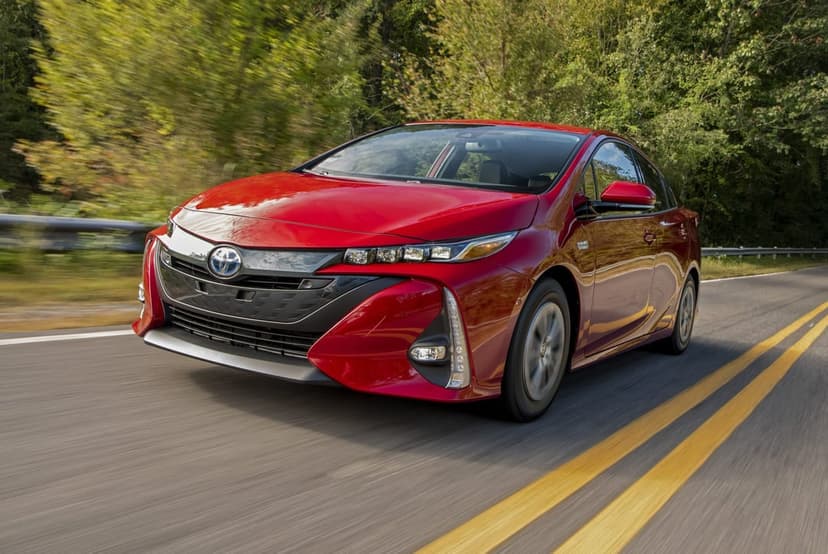 Red Toyota Prius Prime driving on a paved road with motion-blurred trees and double yellow lines.