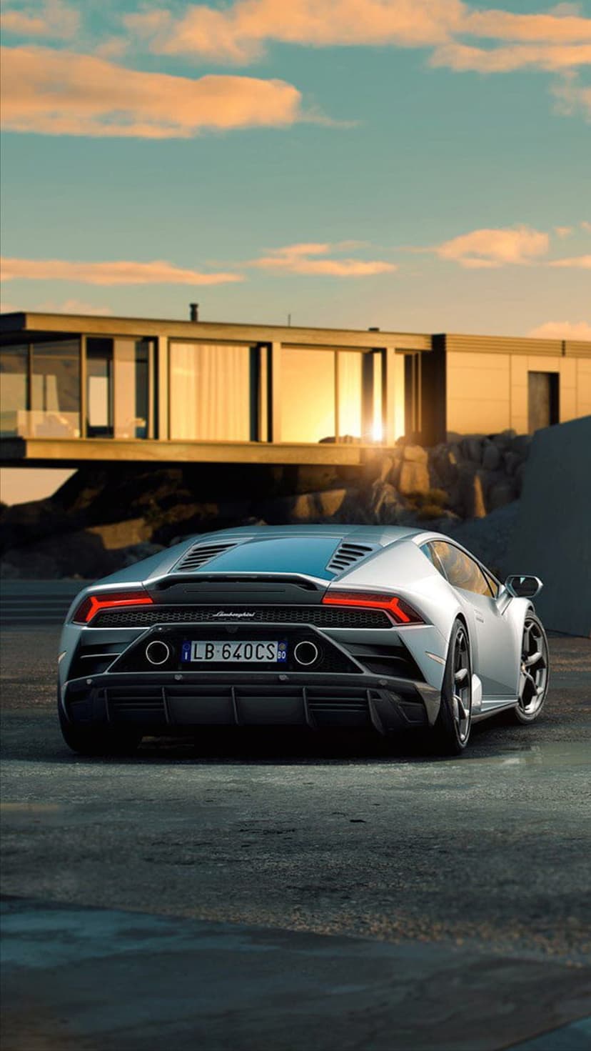 Silver Lamborghini supercar parked outside a modern glass house during a golden orange sunset with red taillights.