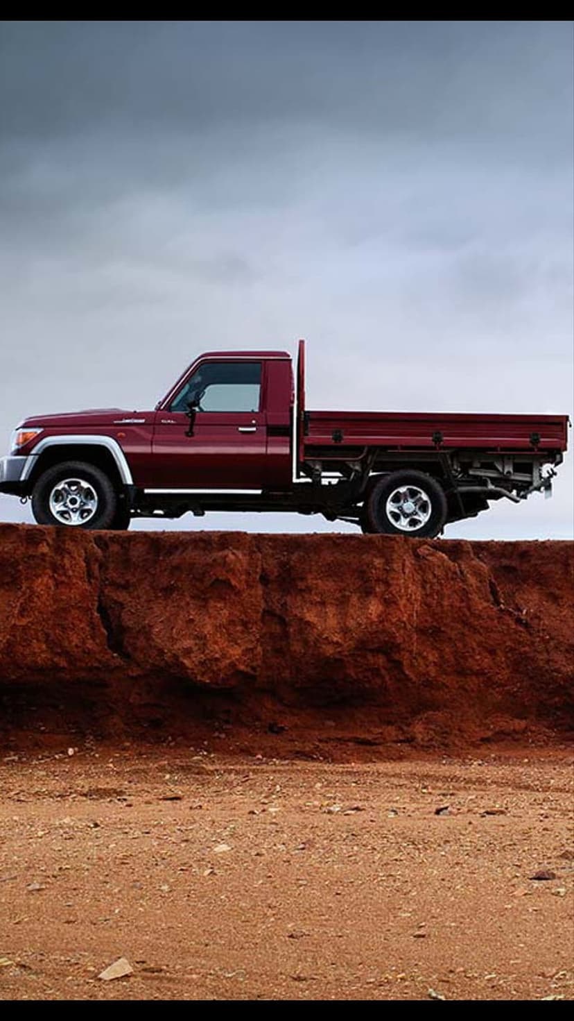 Red Toyota Land Cruiser pickup truck parked on a dusty cliff edge under a moody, overcast sky with rocky ground.