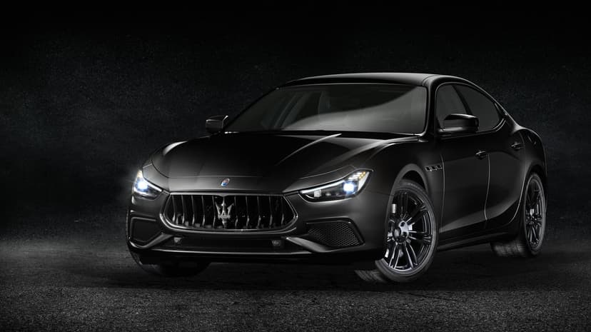 Black Maserati Ghibli sedan with glowing headlights on dark asphalt under dramatic studio lighting.