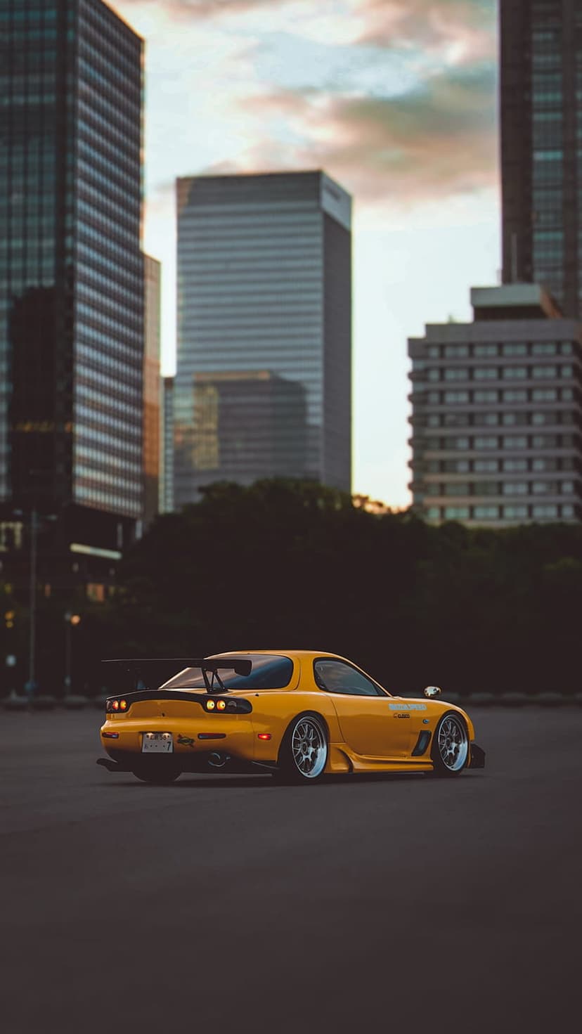 Yellow Mazda RX-7 with a large rear spoiler parked on an urban street against a backdrop of glass skyscrapers.