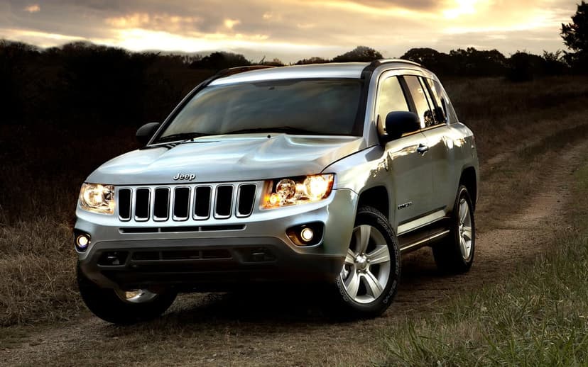 Silver Jeep Compass SUV driving on a dirt trail through a field during a golden hour sunset with lights on.