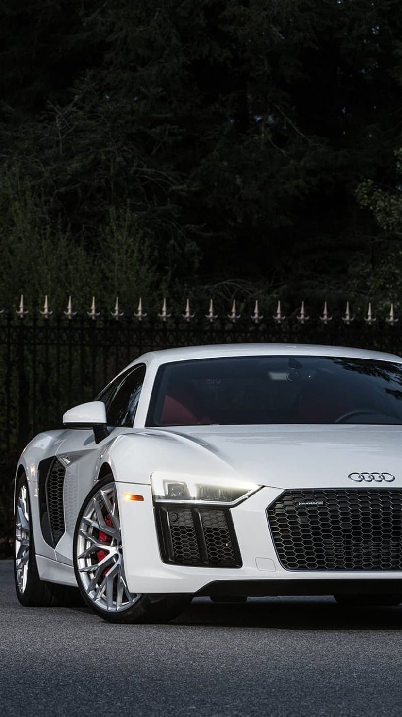 White Audi R8 supercar with red interior and LED headlights parked on asphalt near an ornamental black fence.