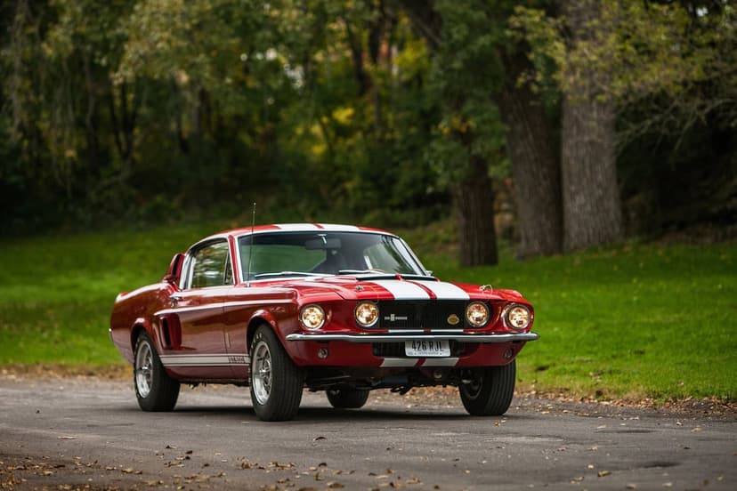 Red 1967 Shelby Mustang GT500 with white stripes and glowing headlights on a tree-lined asphalt road.