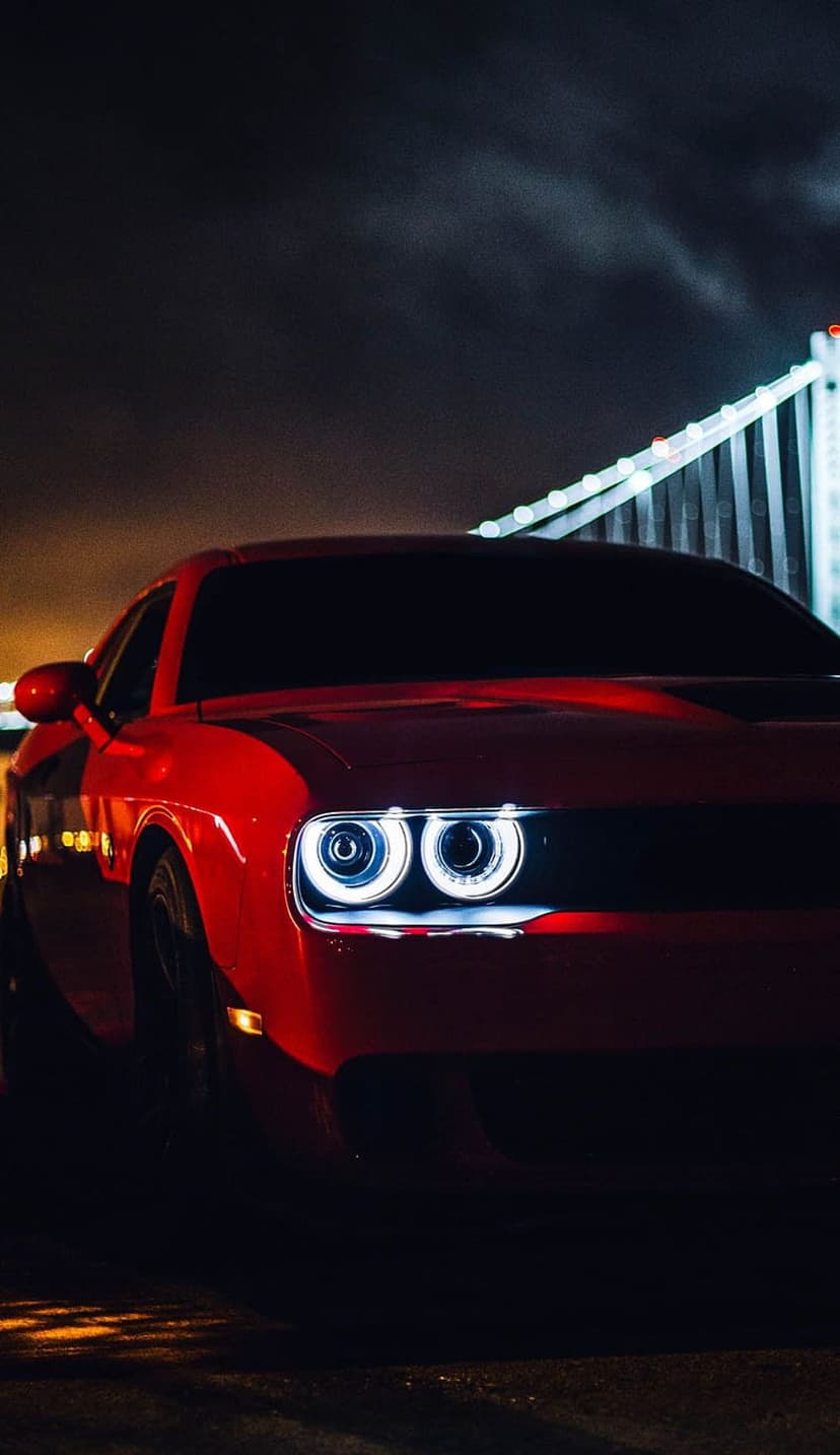 Red Dodge Challenger with luminous dual headlights stands against a bokeh background of city bridge lights.