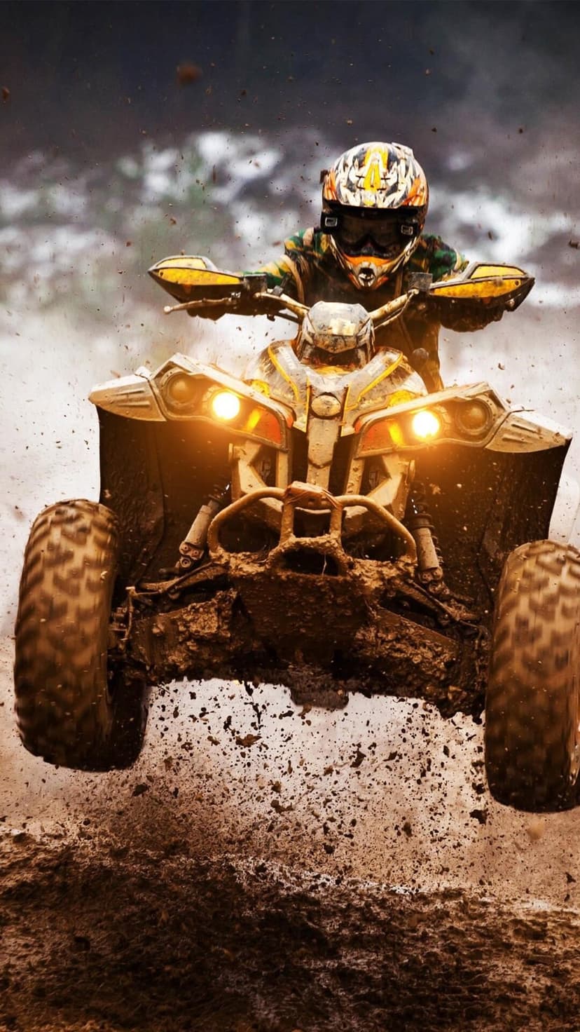 Muddy ATV rider wearing a helmet jumps through water, kicking up a spray of dirt with bright headlights glowing.