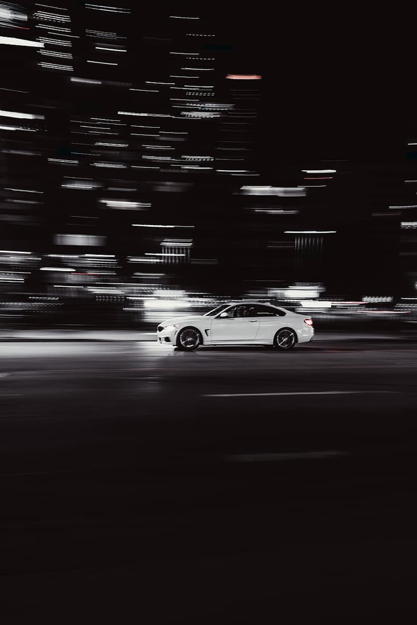 White BMW luxury sedan speeds through urban streets with colorful blurred light trails at nighttime.