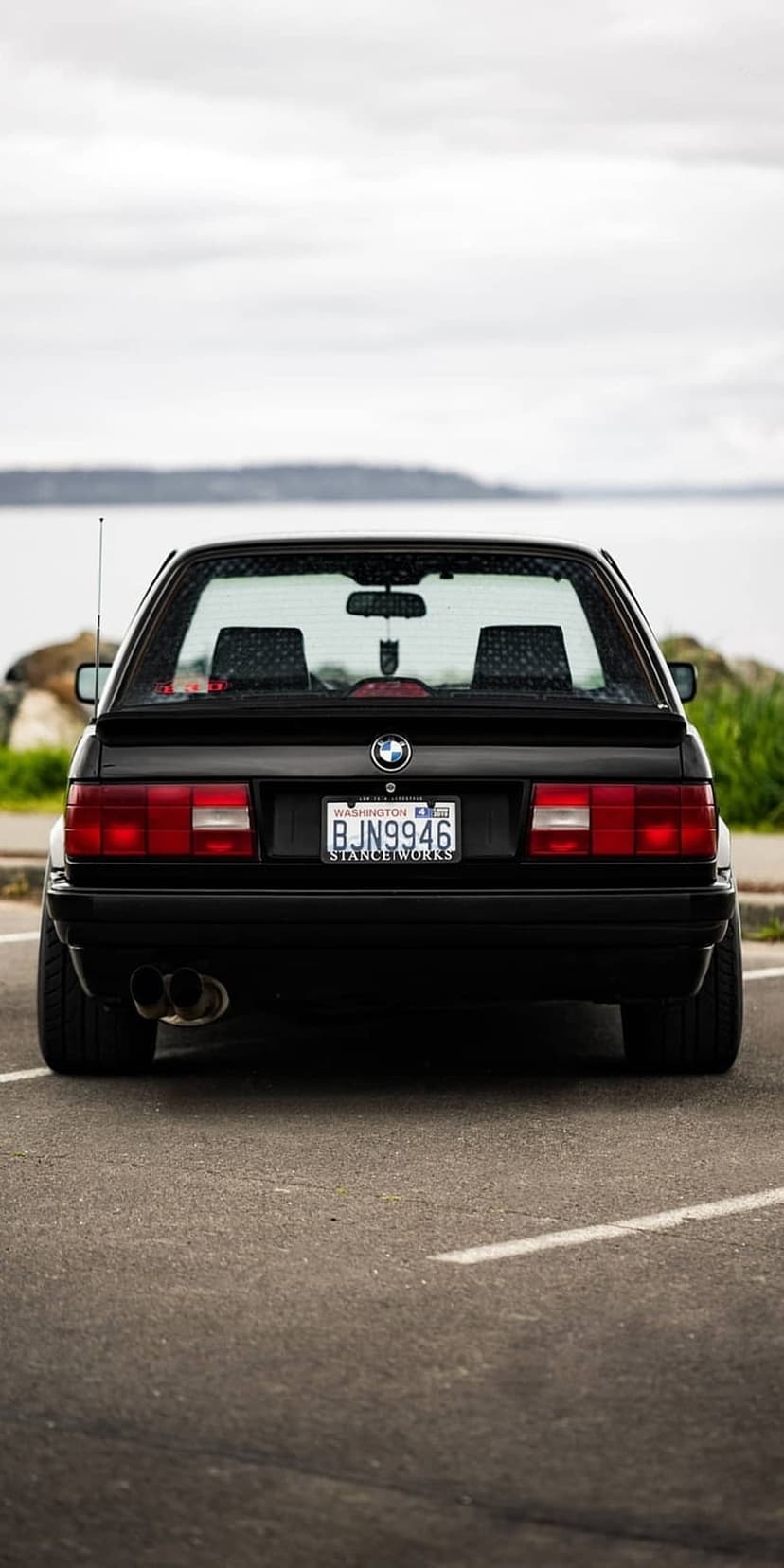 Black BMW E30 rear view showing Washington plates and dual exhausts parked by a serene waterfront under grey clouds.
