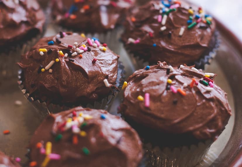 Chocolate cupcakes topped with dark cocoa frosting and rainbow sprinkles are arranged on a light-colored platter.