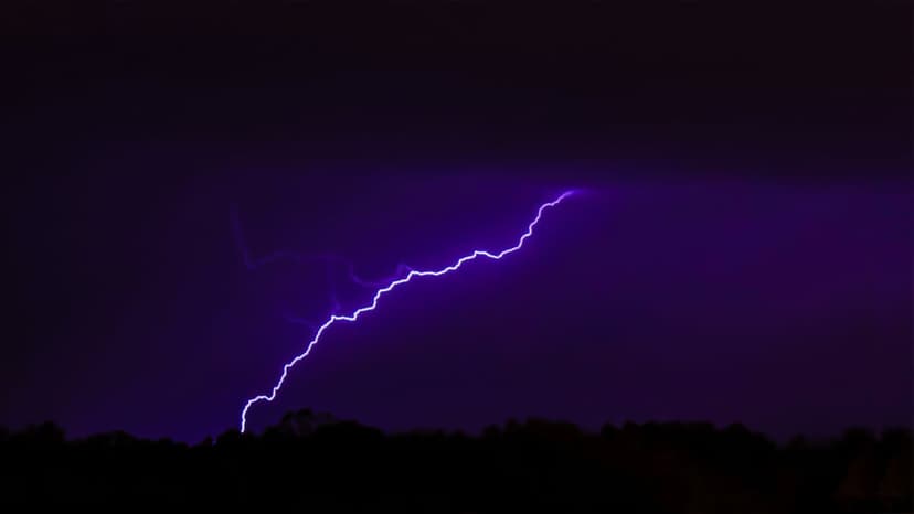 Purple lightning bolt zigzags through dark clouds above a silhouetted horizon during a nighttime thunderstorm.