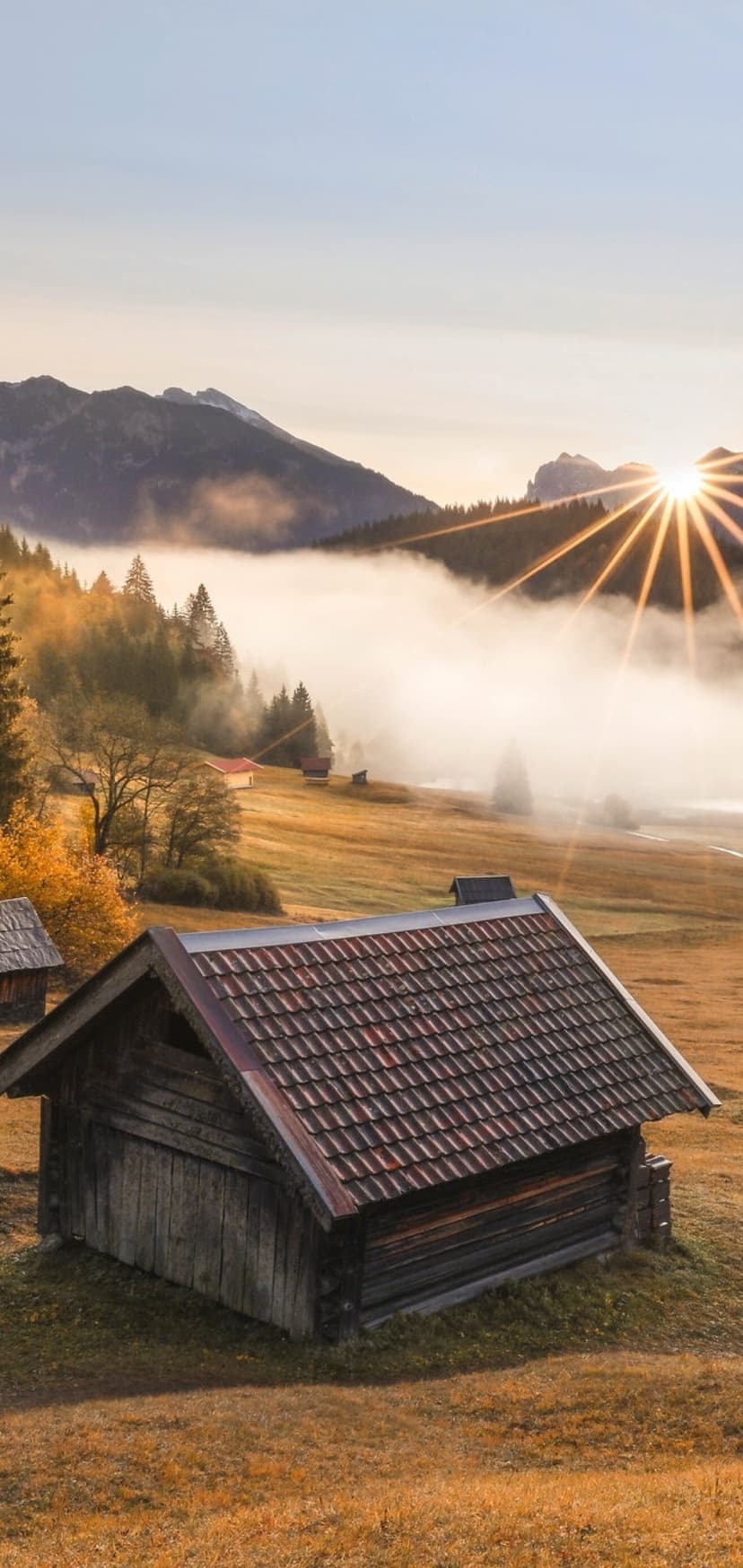 Weathered wooden cabin in a golden field with sun rays hitting misty mountains in a vertical landscape view.