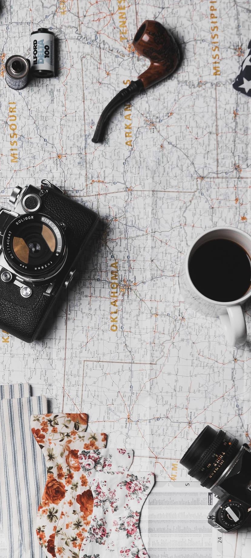 Vintage film camera, wooden pipe, and a cup of coffee rest on a folded paper map in a moody flatlay scene.