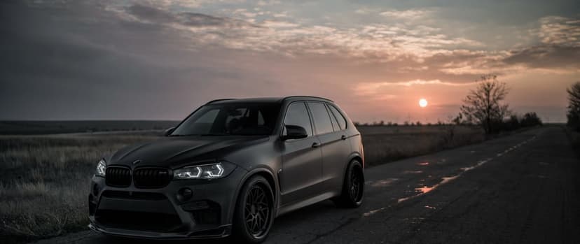 Matte black BMW X5 M on a rural road at sunset with glowing headlights and an orange hazy sky background.