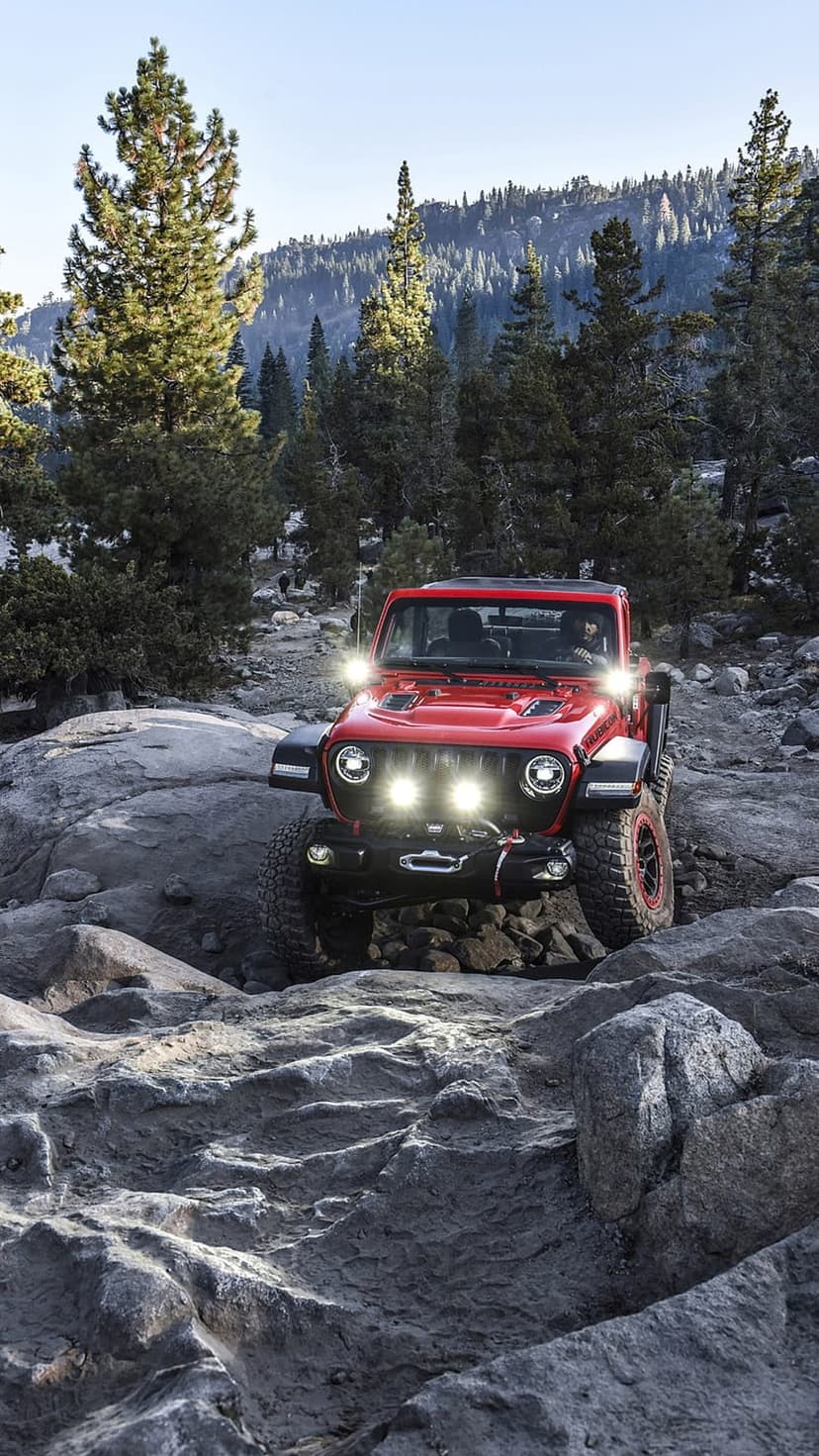 Red Jeep Wrangler navigating a steep rocky slope in a misty pine forest with its headlights glowing bright.