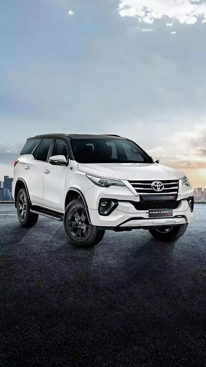 White Toyota Fortuner with black roof and rims parked on asphalt with a city skyline in the background.