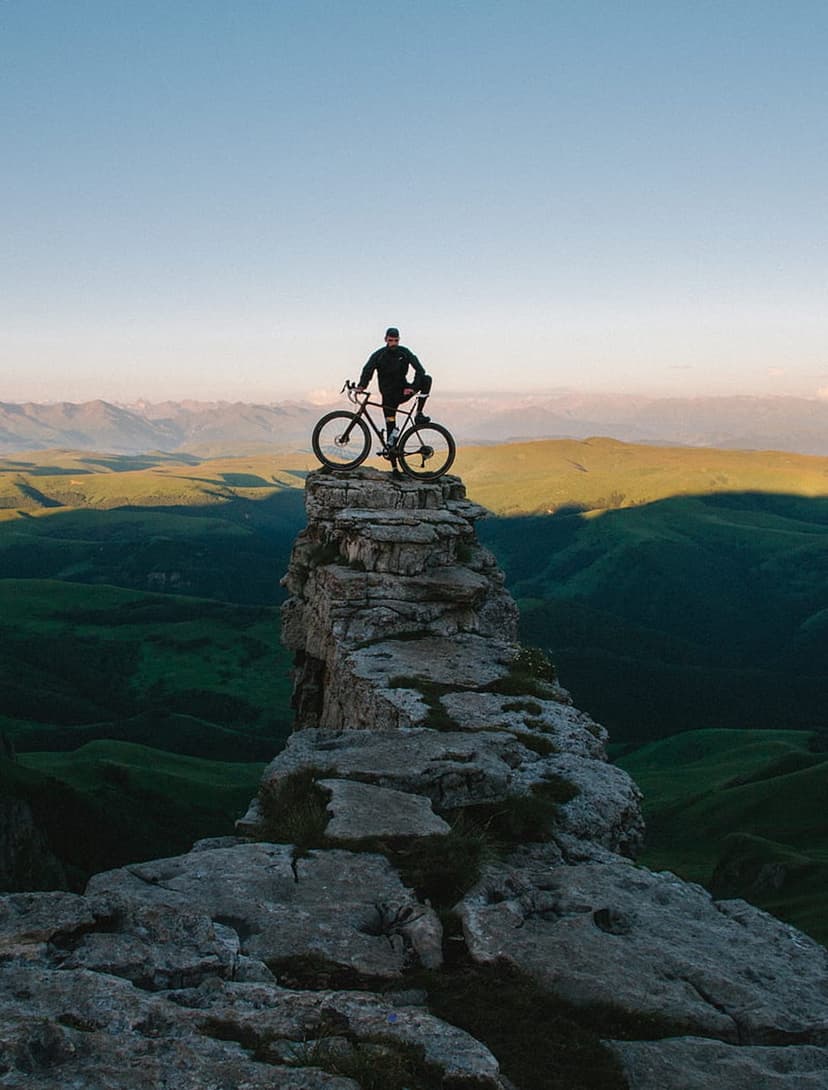 Silhouetted cyclist stands with a bike on a rocky mountain pinnacle overlooking misty green hills at sunrise.