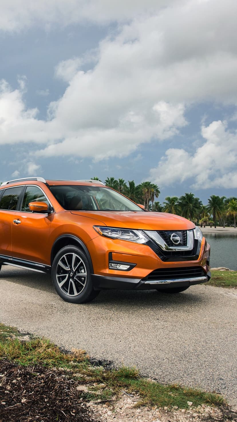 A bright orange SUV parked on a coastal road with lush palm trees and a calm lagoon under a soft cloudy sky.