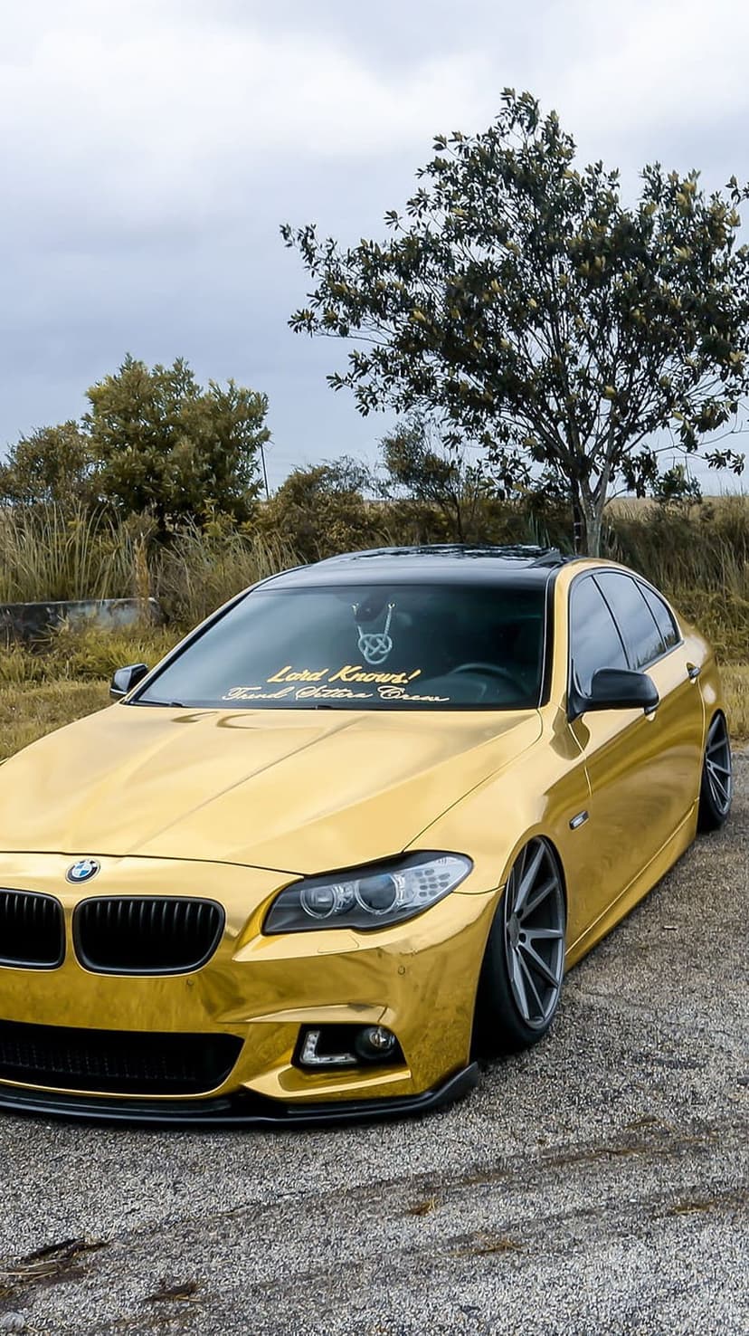 Gold BMW sedan with black custom rims and a lowered stance parked on gravel against a background of trees.
