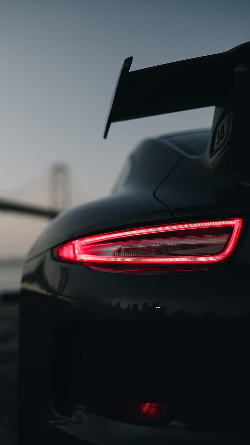 Porsche tail lights glow red on a black sports car with a blurred suspension bridge and city skyline at dusk.